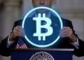 Un hombre sostiene un circulo con el logo hologrรกfico de Bitcoin sobre un podio en el que hay una bandera de Estados Unidos.