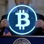 Un hombre sostiene un circulo con el logo holográfico de Bitcoin sobre un podio en el que hay una bandera de Estados Unidos.