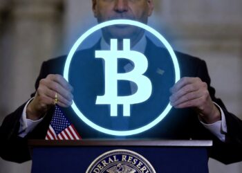 Un hombre sostiene un circulo con el logo holográfico de Bitcoin sobre un podio en el que hay una bandera de Estados Unidos.