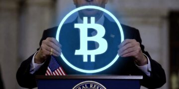 Un hombre sostiene un circulo con el logo holográfico de Bitcoin sobre un podio en el que hay una bandera de Estados Unidos.