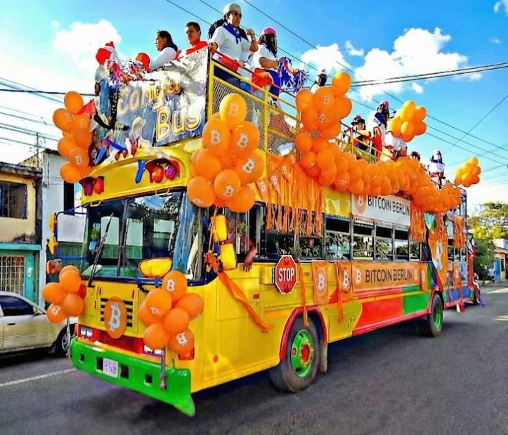Un bus amarillo adoprnado con globos que lucen el logo de bitcoin recorre las calles de Bitcoin Berlín en El Salvador.
