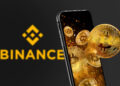 Binance con logo amarillo y un smartphone del que emergen monedas físicas de bitcoin doradas.