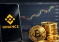 Smartphone con el logo de Binance en pantalla junto a monedas fรญsicas de bitcoin, con grรกficos del mercado de fondo.