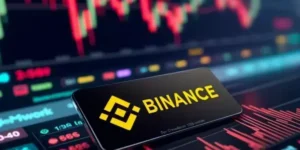 Un celular que muestra el logo de binance en su pantalla frente a indicadores de mercados.