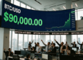 En una oficina moderna y luminosa con vistas a la ciudad, un grupo de corredores de bolsa celebra con los brazos en alto frente a una gigantesca pantalla curva que muestra en verde brillante un gráfico ascendente y el precio de Bitcoin alcanzando los 90.000 dólares.