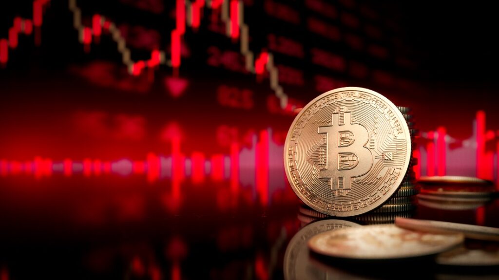Monedas físicas de bitcoin apiladas en primer plano, con un gráfico de velas rojas en el fondo.