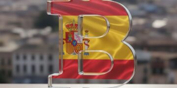 Logo de bitcoin en cristal que muestra la bandera de España y de fondo una ciudad.