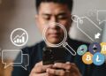 una persona usando el celular, detrás gráficos de precio, monedas de criptomonedas y una lupa observando lo que la gente dice en X.