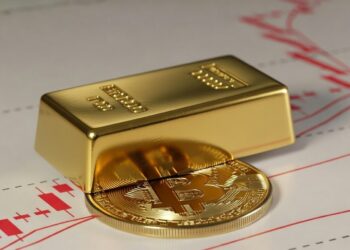 un lingote de oro sobre una moneda de bitcoin y detrás gráficos de precios rojos.