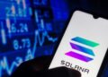 Una mano sujetando un teléfono inteligente que muestra el logo de Solana, y de fondo una pared de gráficas en color azul.