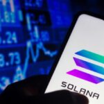 Una mano sujetando un teléfono inteligente que muestra el logo de Solana, y de fondo una pared de gráficas en color azul.