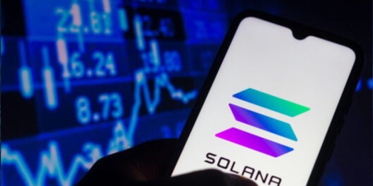 Una mano sujetando un teléfono inteligente que muestra el logo de Solana, y de fondo una pared de gráficas en color azul.