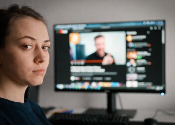 Una mujer joven de expresión aburrida y mirada perdida, en el fondo una pantalla mostrando un canal de YouTube.