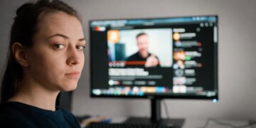 Una mujer joven de expresión aburrida y mirada perdida, en el fondo una pantalla mostrando un canal de YouTube.