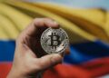 La mano de una persona sostiene una moneda de bitcoin. De fondo se observa la bandera de Colombia.