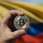 La mano de una persona sostiene una moneda de bitcoin. De fondo se observa la bandera de Colombia.