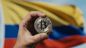 La mano de una persona sostiene una moneda de bitcoin. De fondo se observa la bandera de Colombia.