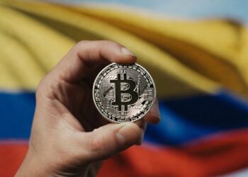La mano de una persona sostiene una moneda de bitcoin. De fondo se observa la bandera de Colombia.