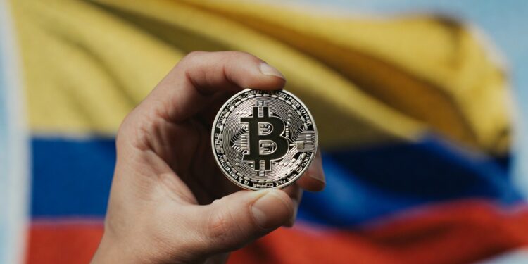 La mano de una persona sostiene una moneda de bitcoin. De fondo se observa la bandera de Colombia.