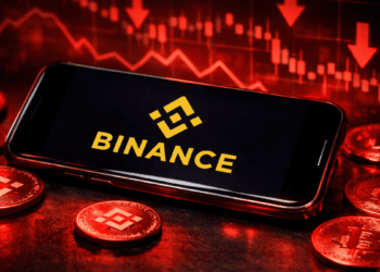 Un celular que muestra el logo de Binance rodeado de monedas físicas de BNB y gráficos de precio rojos.