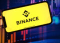 Logo de Binance sobre una pantalla de un móvil con velas rojas y verdes de fondo que reflejan la volatilidad del mercado.