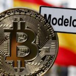 Moneda física de bitcoin dorada con un cartel que dice "Modelo 721" y la bandera de España detrás.