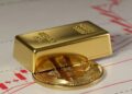 un lingote de oro sobre una moneda de bitcoin y detrás gráficos de precios rojos.