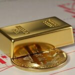 un lingote de oro sobre una moneda de bitcoin y detrás gráficos de precios rojos.