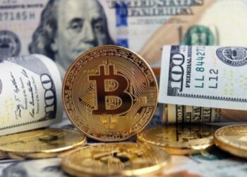 Moneda física de bitcoin dorada rodeada de billetes de 100 dólares estadounidenses.