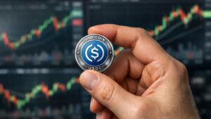 Mano sosteniendo moneda USDC con logo visible frente a grรกficos bursรกtiles desenfocados en fondo financiero dinรกmico.