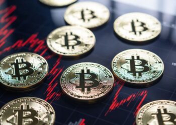 Monedas físicas de bitcoin reposando sobre un gráfico de velas rojo intenso que muestra una fuerte tendencia bajista.