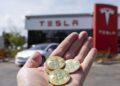 Mano sosteniendo monedas físicas de bitcoin con un concesionario Tesla al fondo.