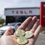 Mano sosteniendo monedas físicas de bitcoin con un concesionario Tesla al fondo.