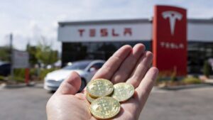 Mano sosteniendo monedas físicas de bitcoin con un concesionario Tesla al fondo.