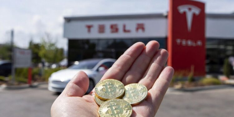Mano sosteniendo monedas físicas de bitcoin con un concesionario Tesla al fondo.