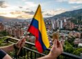 Persona con una bandera de Colombia en la mano izquierda, una moneda física de bitcoin en la derecha y de fondo la ciudad de Medellín.