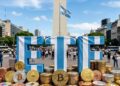 Palabra ETF vestida con un jersey de la selección de Argentina, en el Obelisco. Con monedas físicas de bitcoin y otras criptomonedas apiladas alrededor.