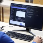 Vista desde atrรกs de una persona con camisa azul sentada frente a un monitor de computadora de escritorio.