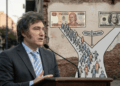 Esta imagen horizontal muestra al presidente de Argentina, Javier Milei, en primer plano con un retrato realista, hablando seriamente detrás de un podio con un micrófono. Viste un traje oscuro, camisa clara y una corbata a rayas celestes y blancas. Detrás de él, sobre una pared de ladrillos, aparece una ilustración esquemática que representa un camino que se bifurca en forma de "Y". En el centro del cruce, un cartel indica hacia la izquierda "PESOS" y hacia la derecha "DÓLARES". El camino de la izquierda lleva a un billete grande de 1000 pesos argentinos, hacia el cual se dirige una multitud de figuras humanas esquematizadas. En contraste, el camino de la derecha, que conduce a un billete de 100 dólares estadounidenses, está completamente vacío, sin ninguna persona.