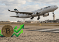 En primer plano, una moneda física de bitcoin con flechas verdes ascendentes. En el fondo, un avión despegando, con bandera iraní.
