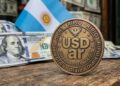 Moneda que tiene grabada la palabra USDar y en los bordes que diga "RIPIO". Con dólares y la bandera de Argentina en el fondo.