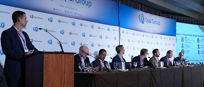 Panel de expertos sentados en una mesa larga durante una conferencia de Opal Group, con un ponente en el podio a la izquierda y un gran cartel corporativo azul y blanco de fondo.