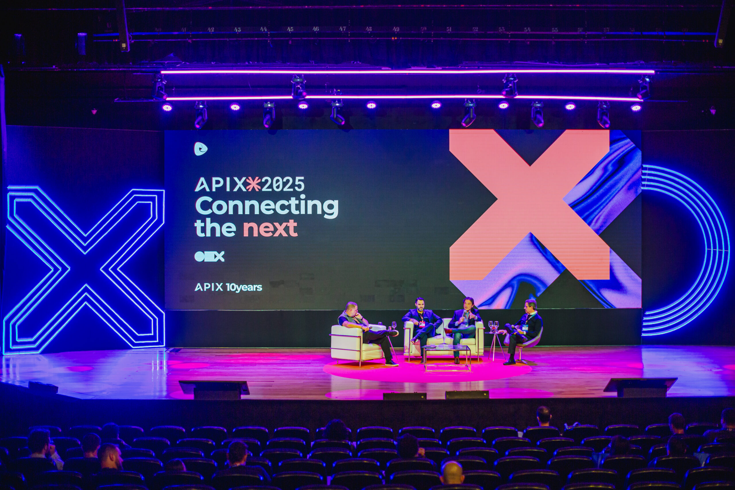 Escenario de conferencia con panelistas, pantalla gigante con texto e iluminación neón.