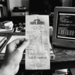 Fotografía histórica en blanco y negro que muestra una mano sosteniendo en primer plano un recibo de Papa John's fechado el 22 de mayo de 2010 con la inscripción "10,000 BTC transferred". Al fondo, sobre una mesa de madera rústica, se observan cajas de pizza y un monitor CRT antiguo de computadora de escritorio.