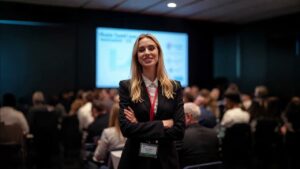 Mujer rubia en traje sonriendo en una conferencia de blockchain con público al fondo.