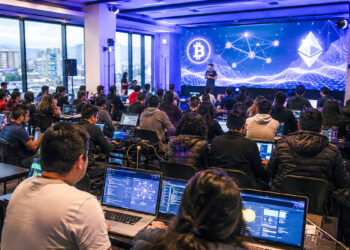 Fotografía de un evento de criptomonedas con una pantalla gigante detras, y todos los concurrentes con notenooks en sus mesas