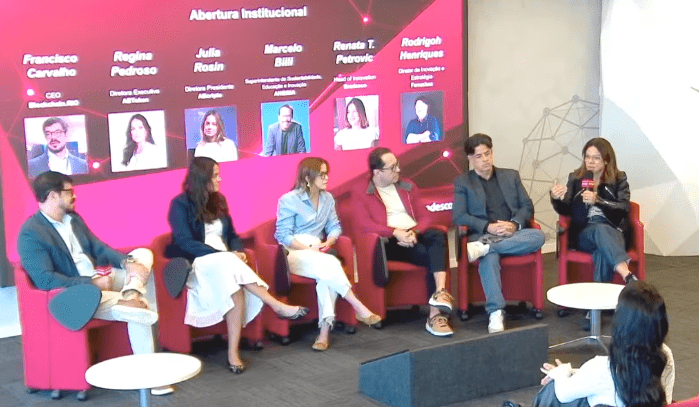 A panel of six finance and technology experts debate sitting on red armchairs in front of an interactive screen with their names. Among them is Renata Petrovic, head of innovation at Bradesco. The event is focused on institutional openness and blockchain technology in Brazil.