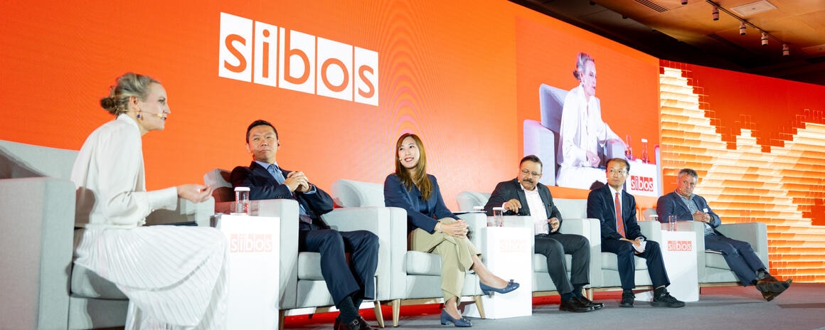 Panelistas en un escenario debatiendo en el evento financiero "Sibos" con fondo color naranja.