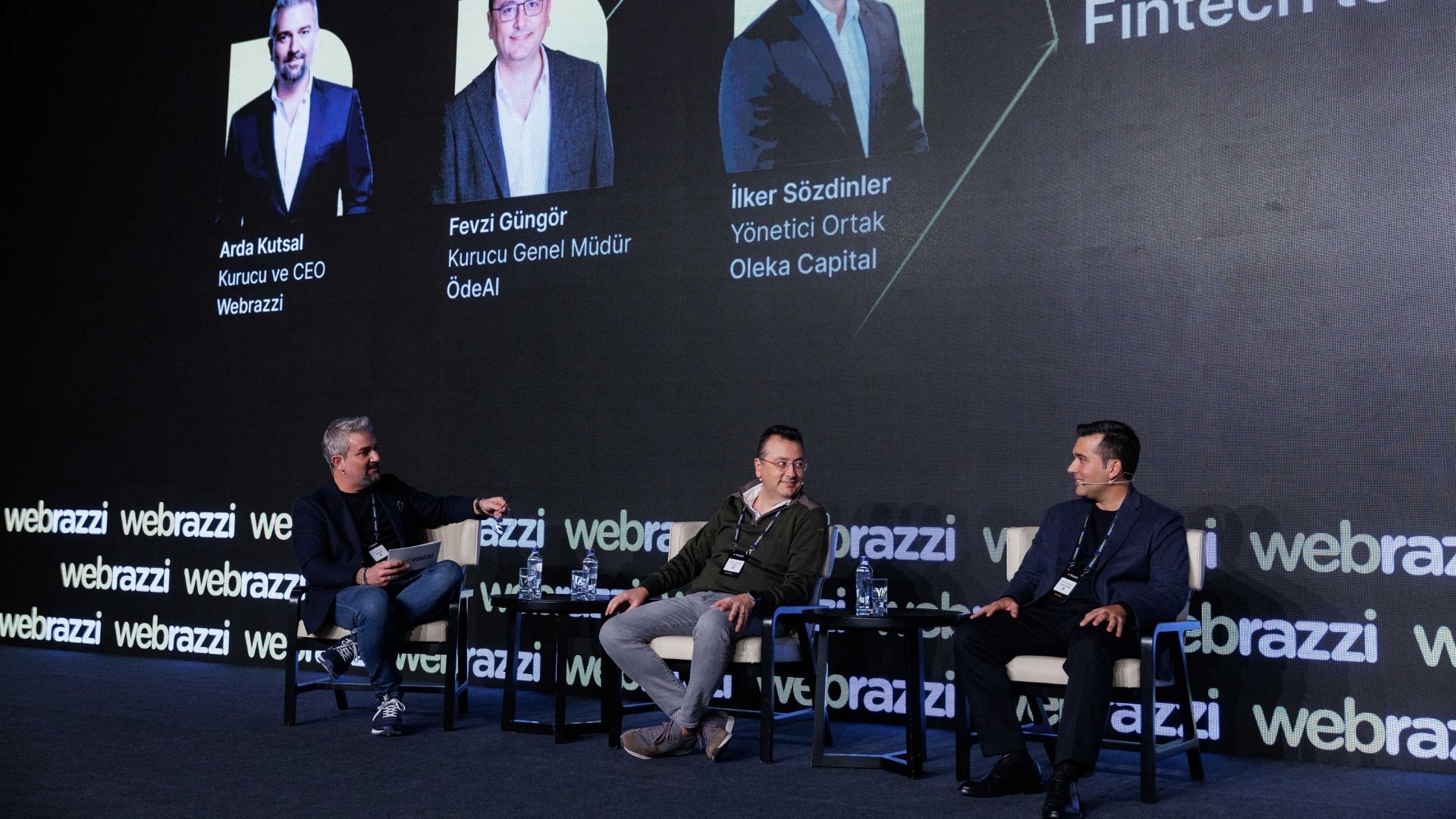 ### Texto Alternativo (Alt Text) > Ponentes conversando sentados en un escenario oscuro con pantalla digital al fondo en Webrazzi Fintech.