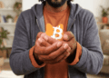 Primer plano de un hombre con vestimenta casual sosteniendo con ambas manos una pequeña alcancía naranja con el logotipo de Bitcoin, simbolizando la protección y la autocustodia de activos digitales en un entorno doméstico.